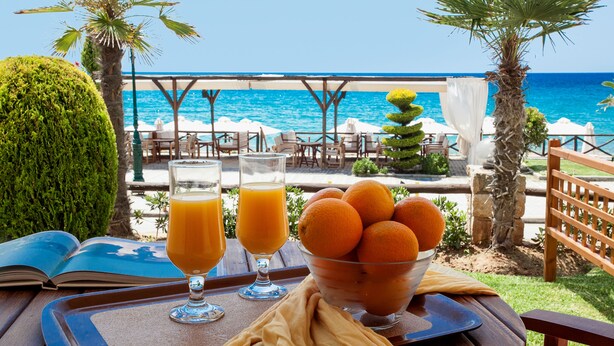 A table with a book, two glasses of orange juice, and a bowl of oranges, set against a backdrop of a beach and umbrellas.