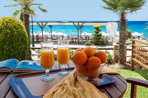 A table with a book, two glasses of orange juice, and a bowl of oranges, set against a backdrop of a beach and umbrellas. - Paralio Hotel (Kassandra)