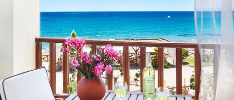 A wooden table with a potted plant, a glass of white wine, and a fruit platter on a balcony overlooking the sea.