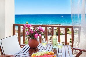 Une table en bois, ornée d’un pot de fleurs, d’un verre de vin blanc et d’une assiette de fruits, est placée sur un balcon donnant sur la mer.