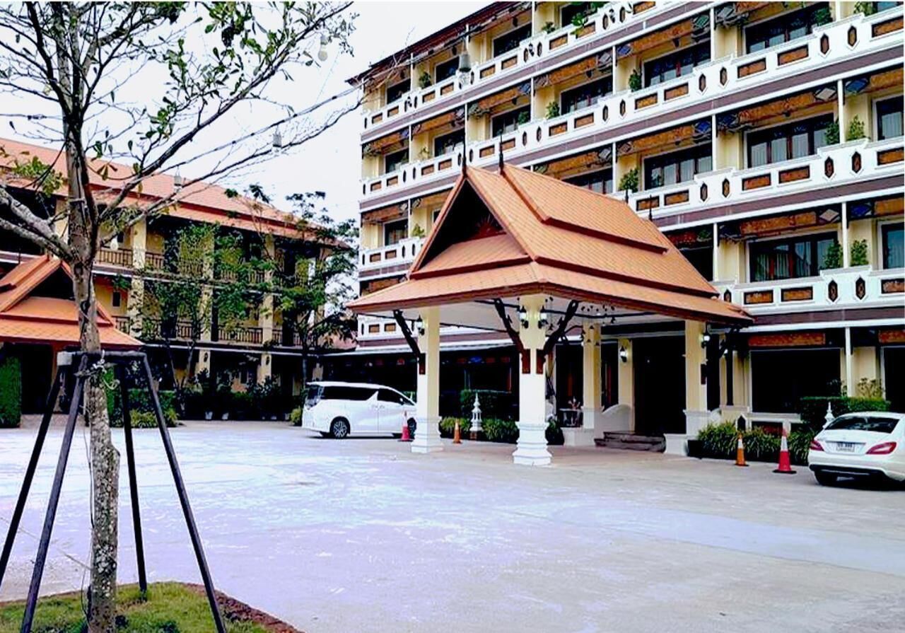 Un hotel de varios pisos con un patio central, coches blancos estacionados al frente y una zona de estar cubierta con un árbol en primer plano.