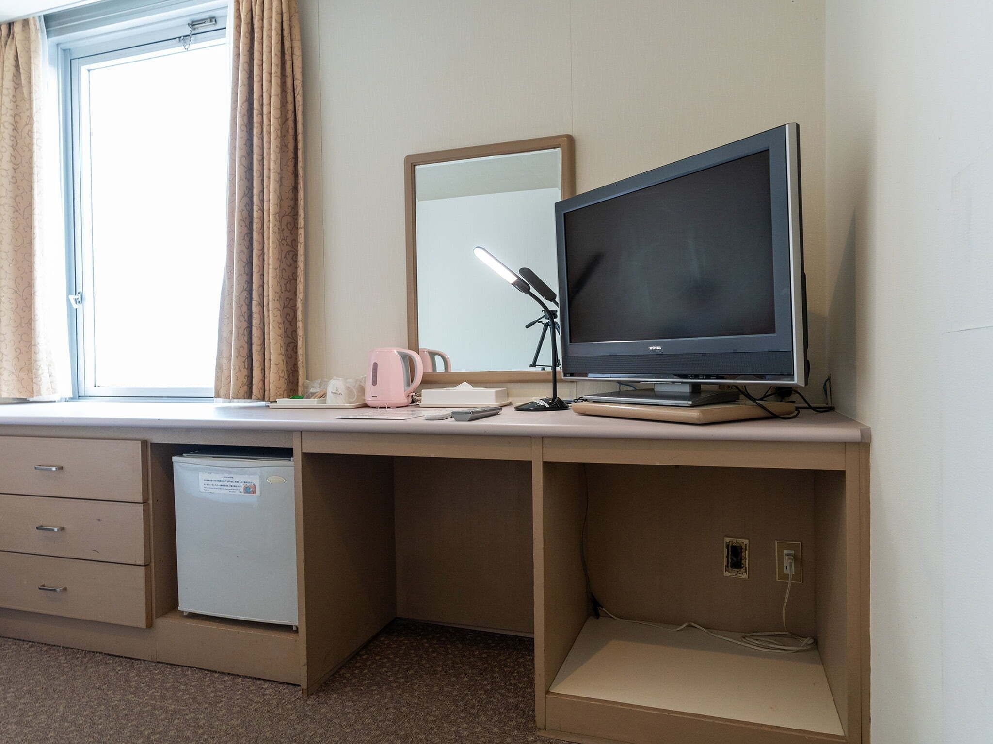 A hotel room with a desk, a small refrigerator, a mirror, a television, and a window with curtains.