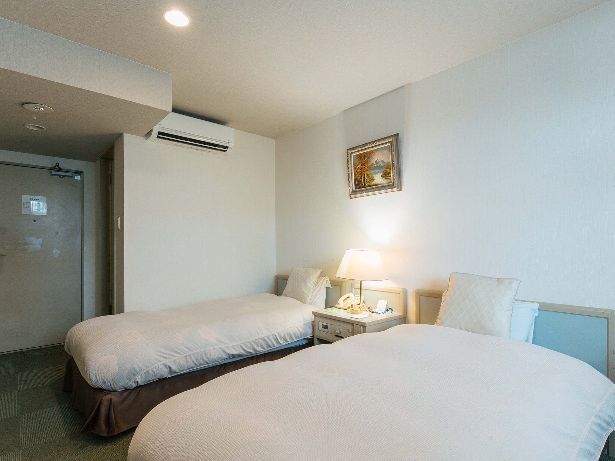 Habitación de hotel con dos camas, una mesita de noche con lámpara, un equipo de aire acondicionado y una puerta.