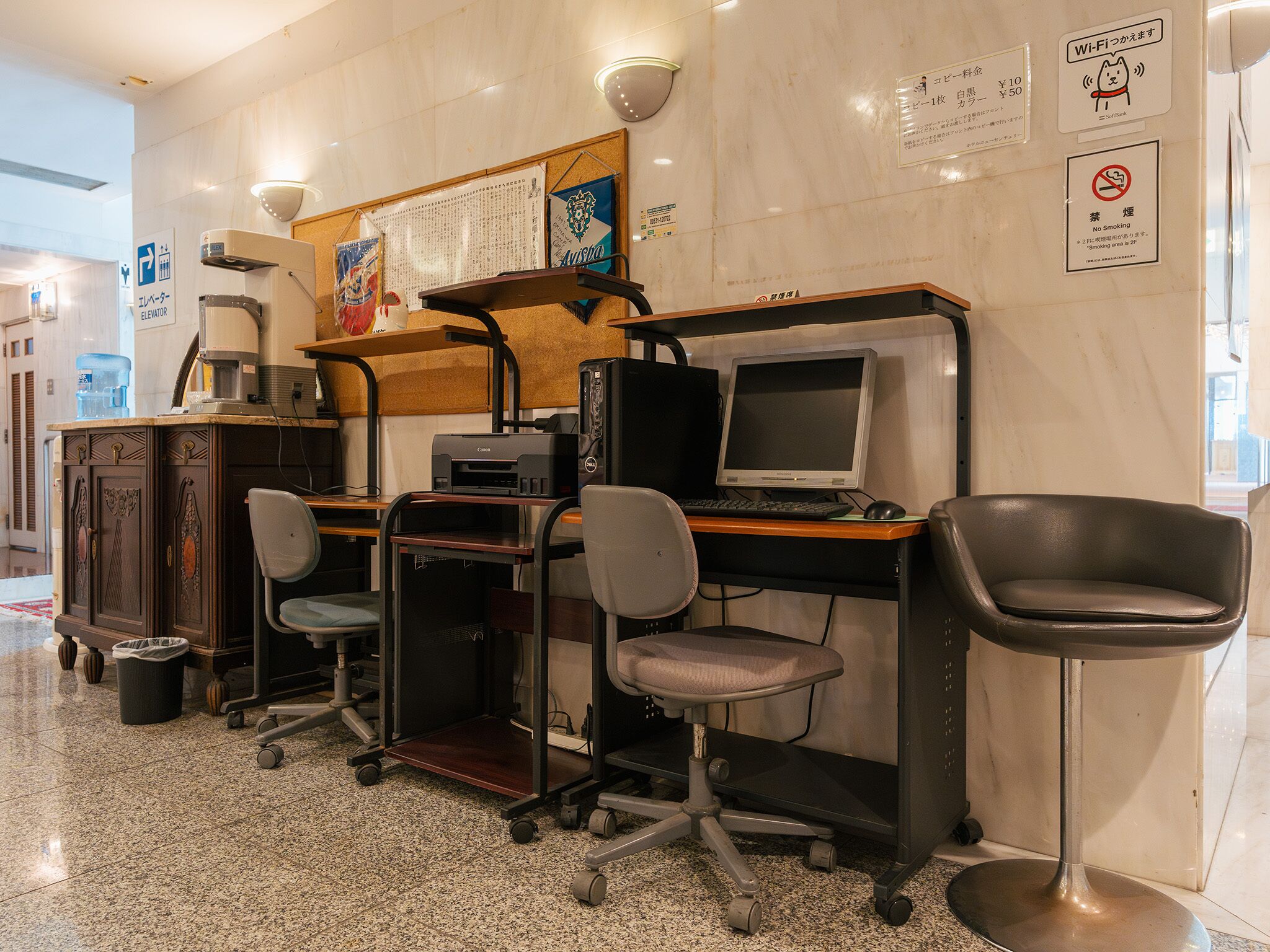 A workspace with a computer, chairs, and a coffee machine. There are signs on the wall indicating amenities and rules.