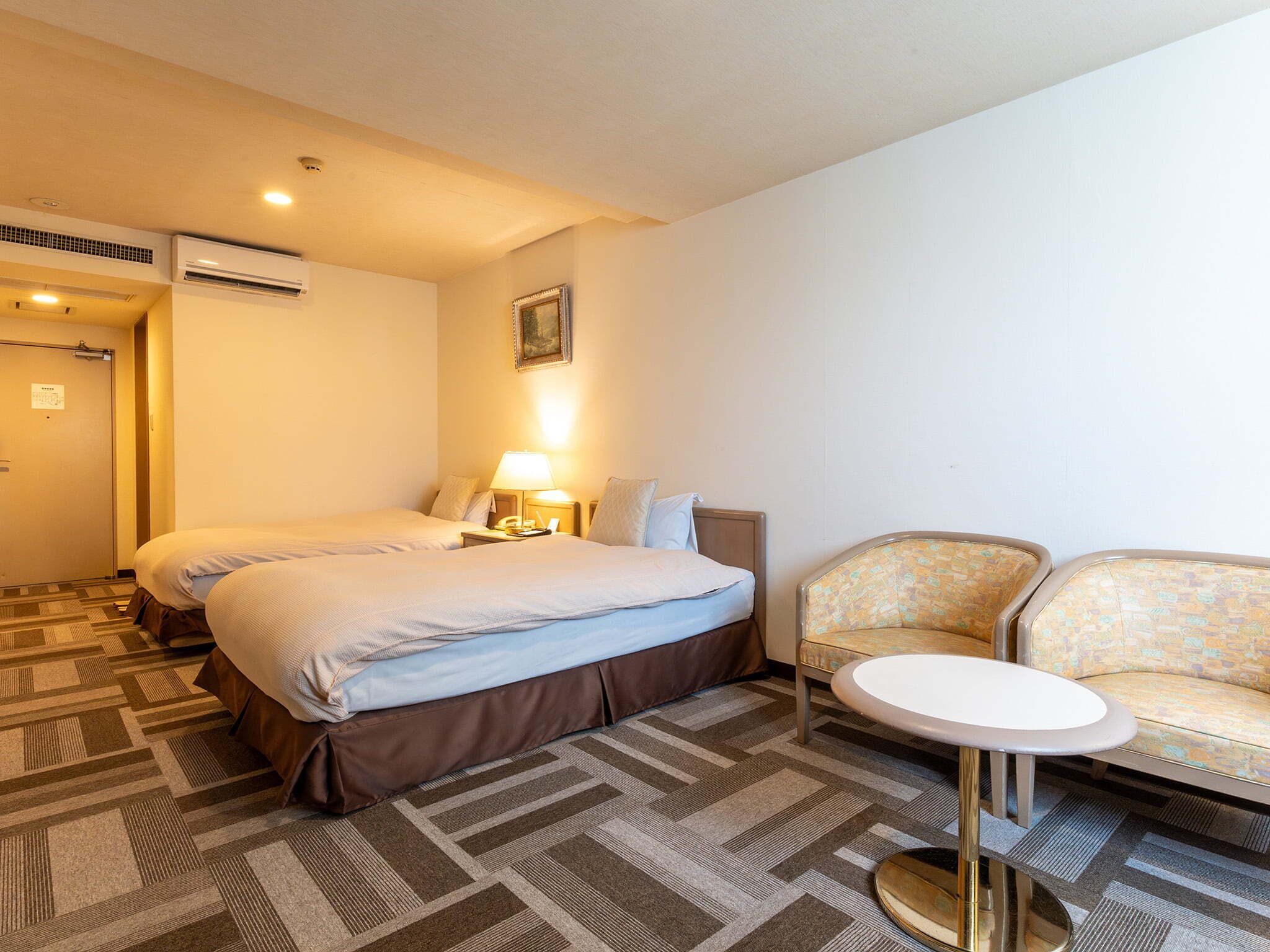 Quarto de hotel com duas camas, um sofá, uma mesinha e ar condicionado fixado na parede.