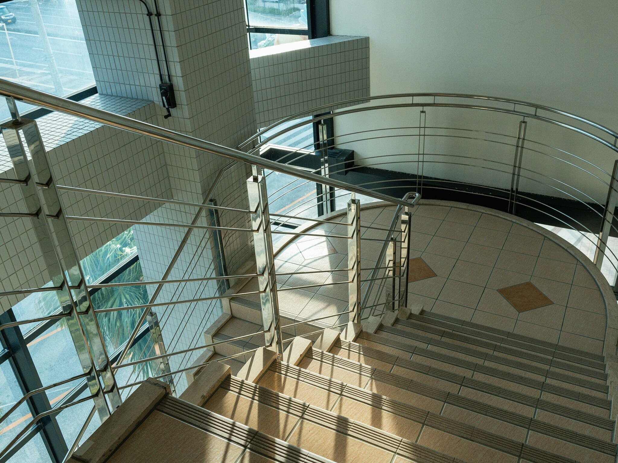 Un escalier en colimaçon avec des rampes en métal et des marches de couleur beige, entouré de murs recouverts de carreaux blancs et d’une fenêtre en verre.