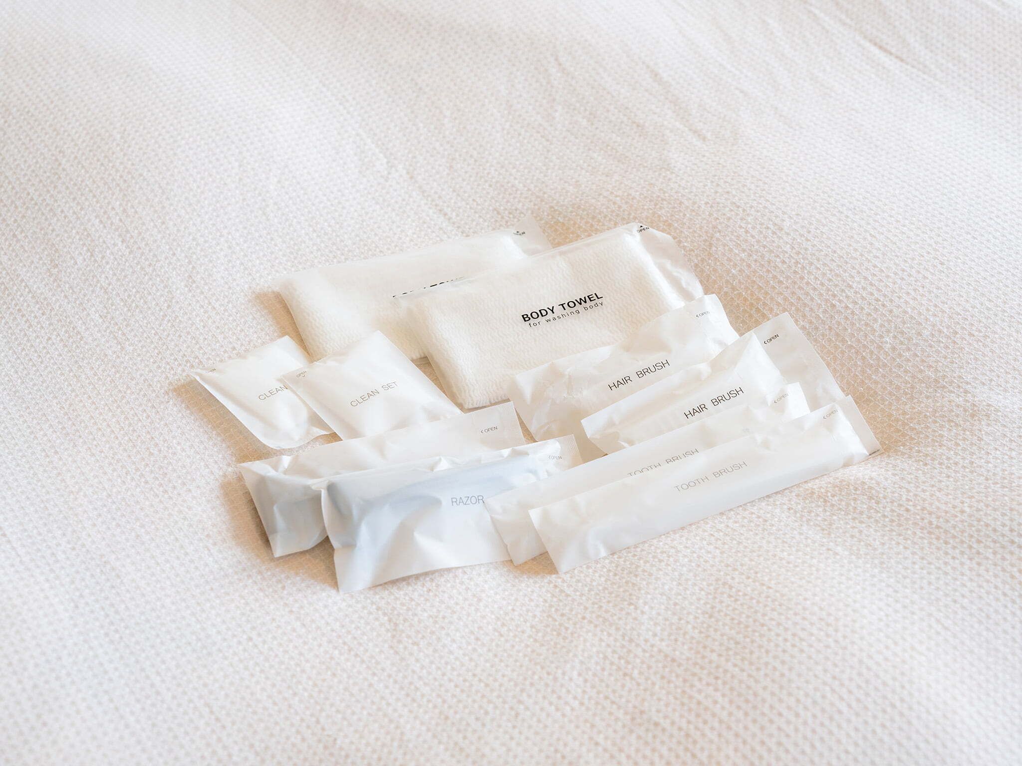 Un conjunto de artículos de tocador blancos, que incluye una toalla de baño, un cepillo para el cabello y un cepillo de dientes, ordenados sobre una superficie blanca con textura.
