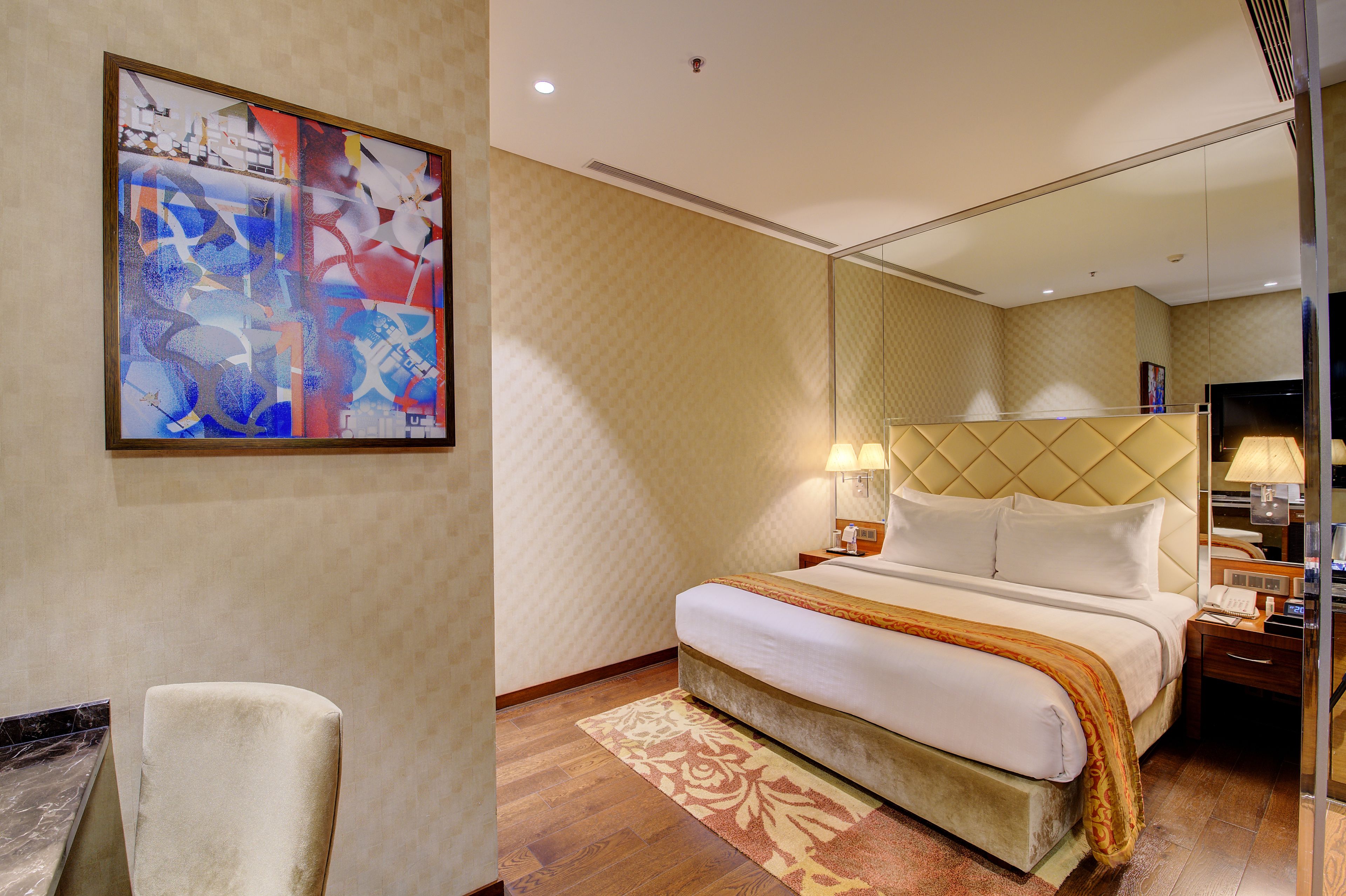 Una habitación de hotel con cama, mesitas de noche, lámpara, una silla y un cuadro abstracto grande en la pared.