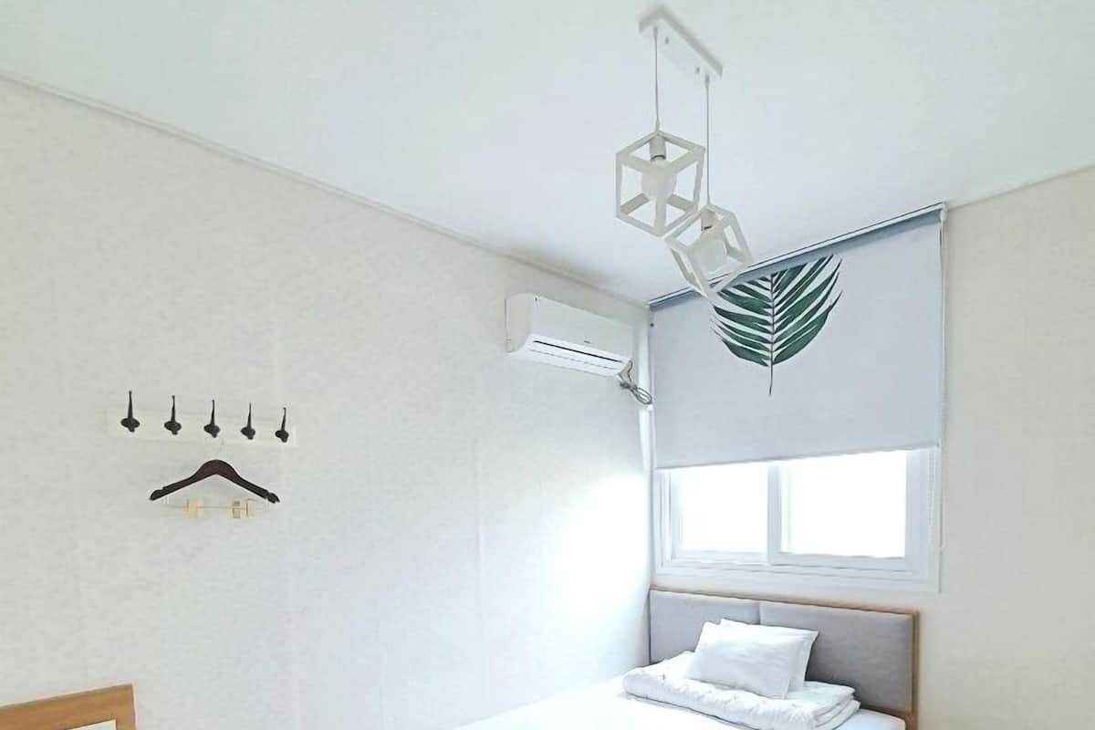 A bedroom with a bed, a wooden nightstand, a hanging light fixture, an air conditioning unit, and a window with a leaf decoration.