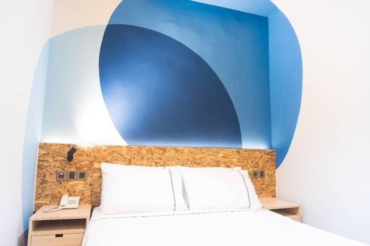 Una habitación de hotel moderna con cabecera de madera, cama con ropa blanca y teléfono montado en la pared.