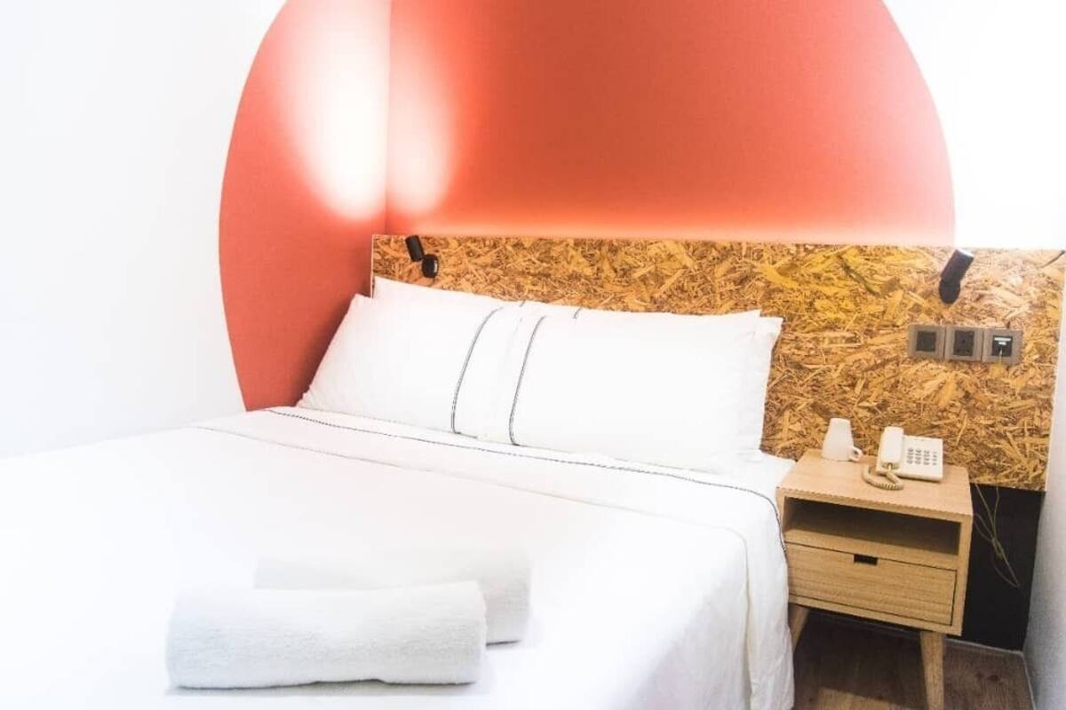 Una cama bien hecha con sábanas blancas y una toalla doblada, una mesita de noche de madera con un teléfono, y una pared con un tablón de corcho.