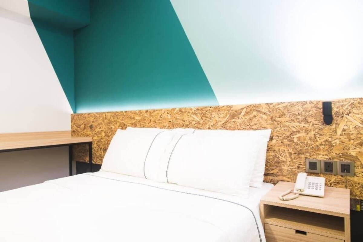 Una habitación de hotel con una cama, cabecera de madera, mesita de noche con un teléfono y un enchufe en la pared.