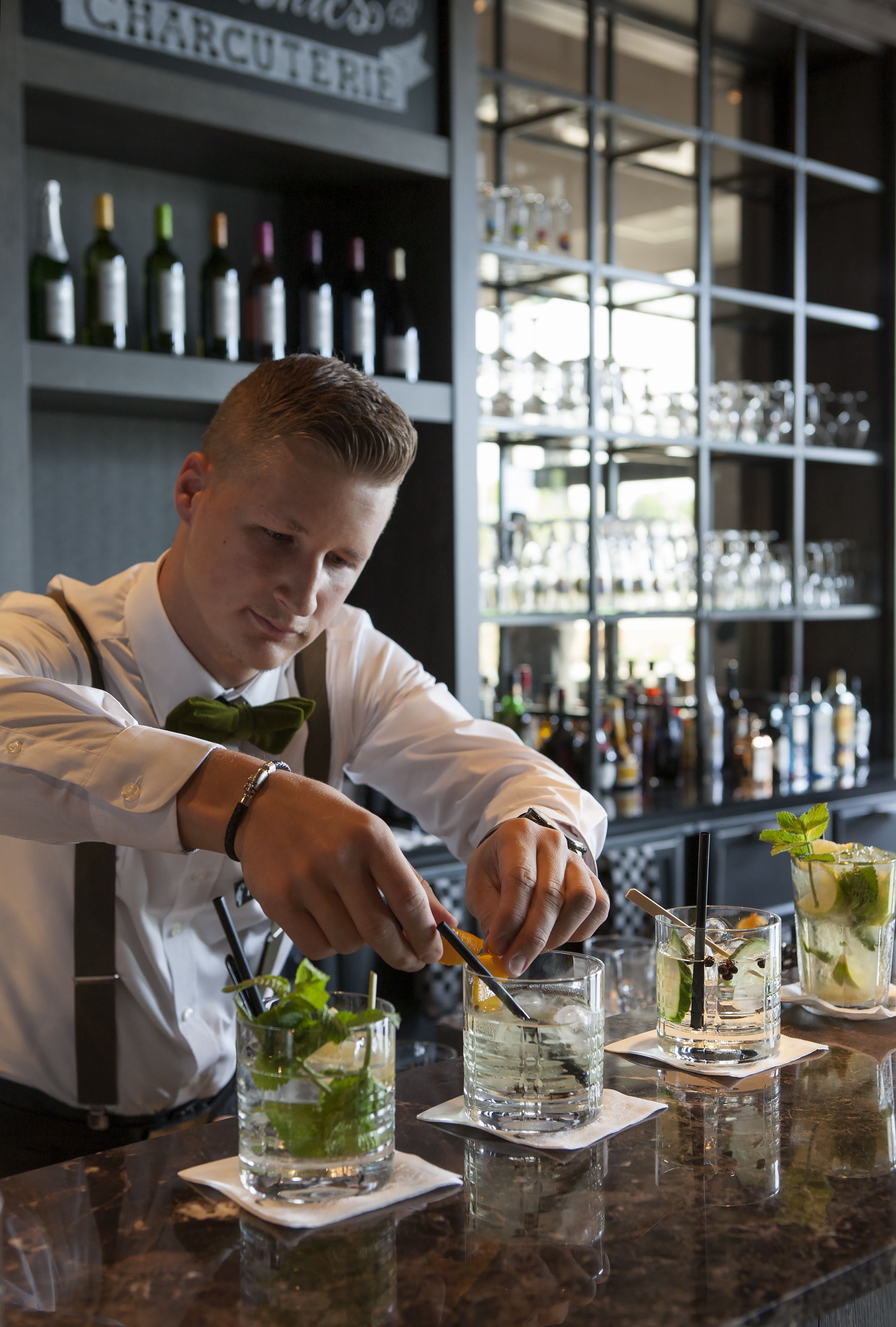 A bartender preparing drinks at a bar with shelves of bottles and glasses in the background.
