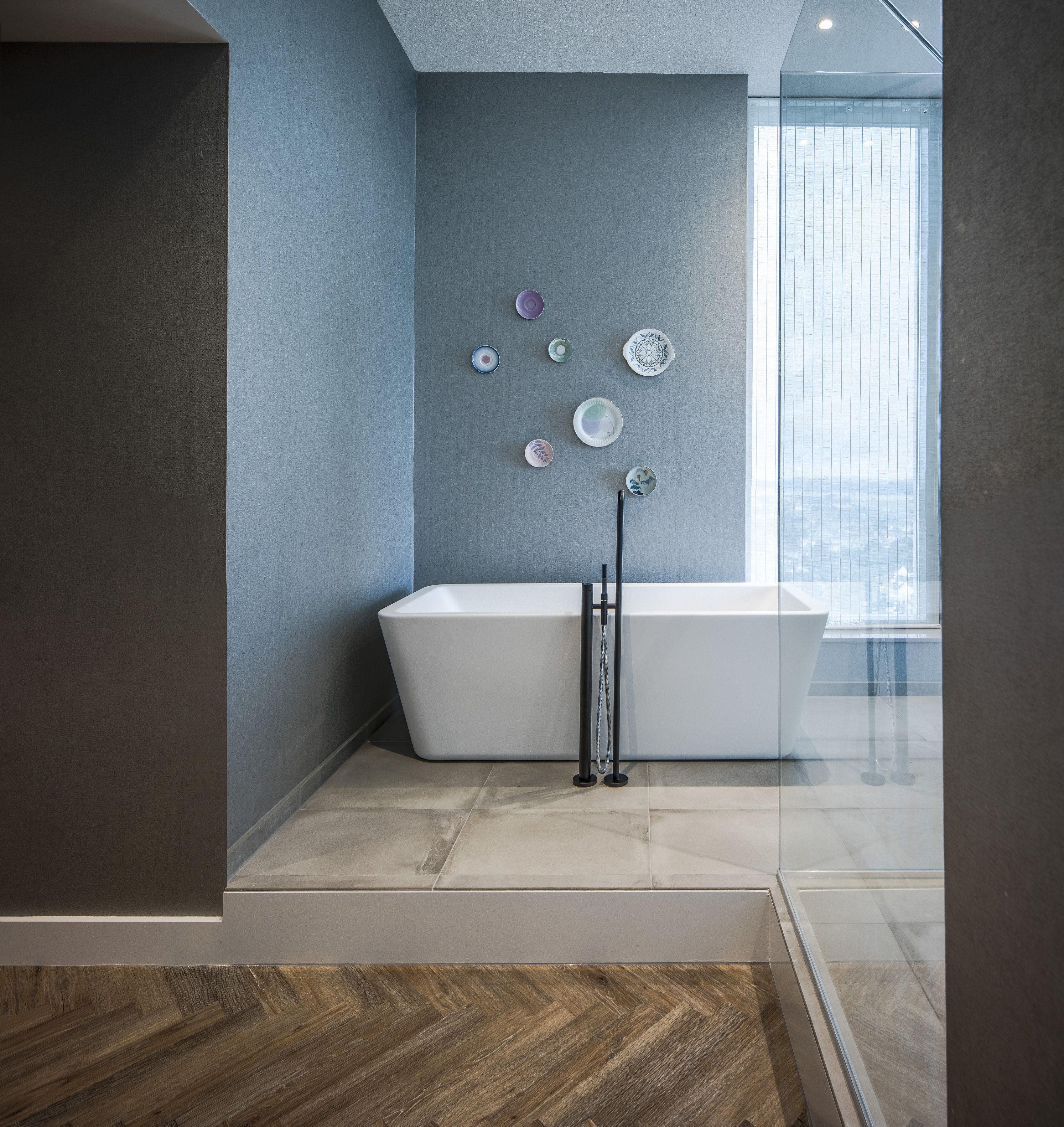 A modern bathroom with a freestanding bathtub, a glass shower enclosure, and a herringbone-patterned wooden floor.