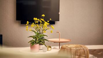 A modern living room with a flat-screen TV mounted on the wall, a round coffee table with a woven stool, and a potted plant with yellow flowers.