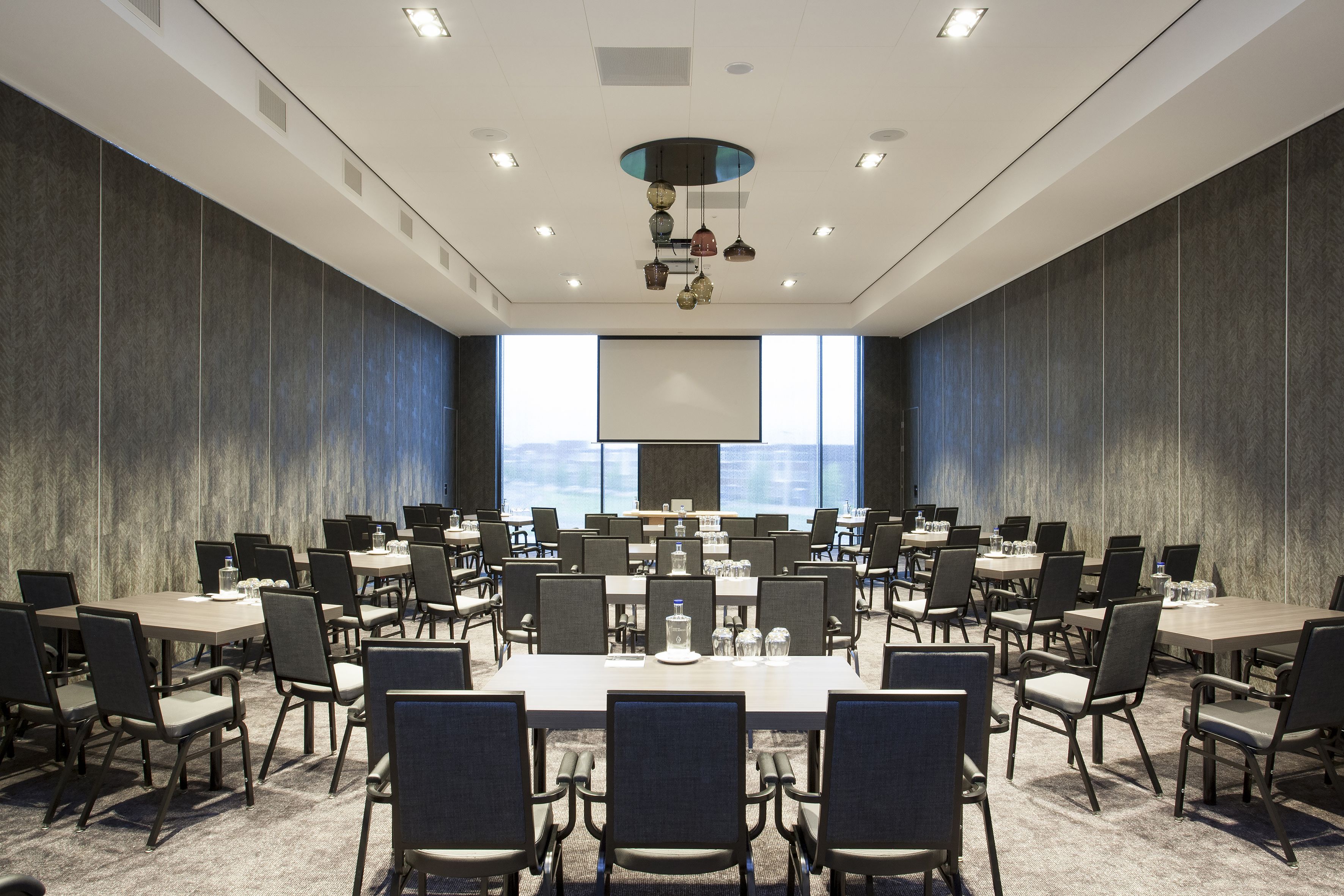 A conference room with multiple tables and chairs, a large screen, and a view of the cityscape through large windows.