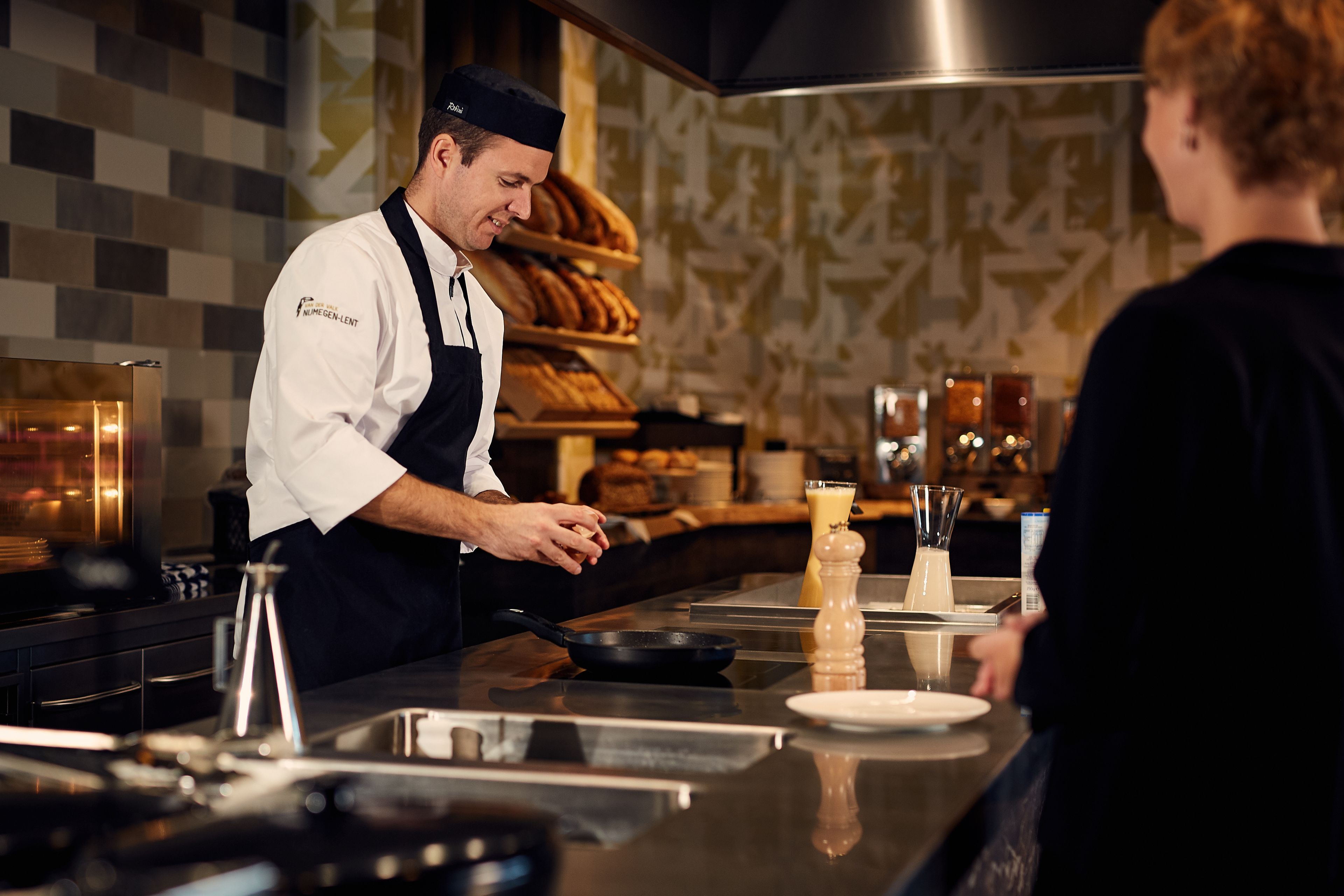 Un chef con uniforme blanco y delantal negro preparando comida en una barra, con una cocina moderna que incluye una sartén, un vaso de leche y un plato.