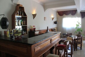 A bar area with a wooden counter, shelves with liquor bottles, and a seating area with wooden chairs and a table. - K.méléon Hôtel (Antananarivo)