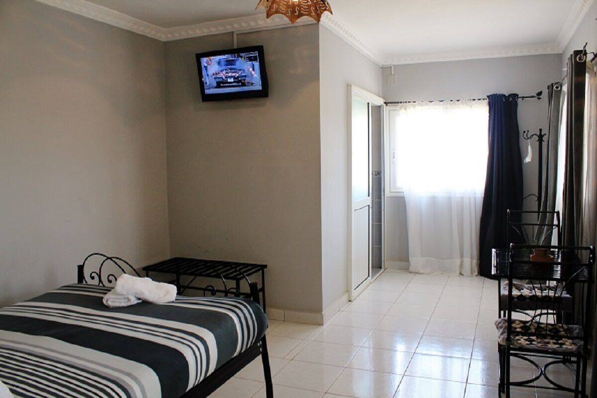 A hotel room with a bed, a small table, a TV mounted on the wall, and a view of the outside through a window with curtains.