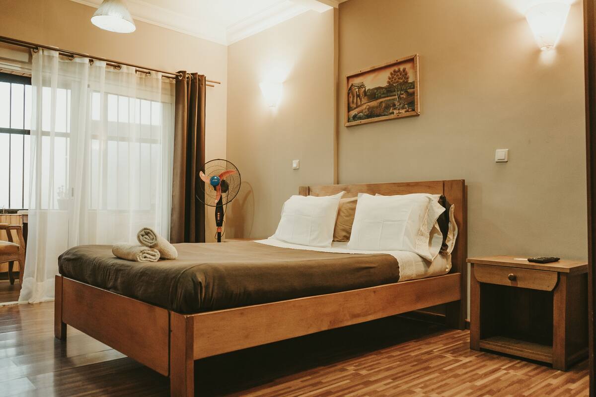 A neatly made bed with a wooden frame, a bedside table with a drawer, and a wall-mounted painting.