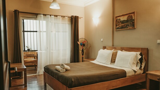 Ein ordentlich eingerichtetes Hotelzimmer mit einem hölzernen Bett, einem Nachttisch, einem Sessel und einem Fenster mit Vorhängen.