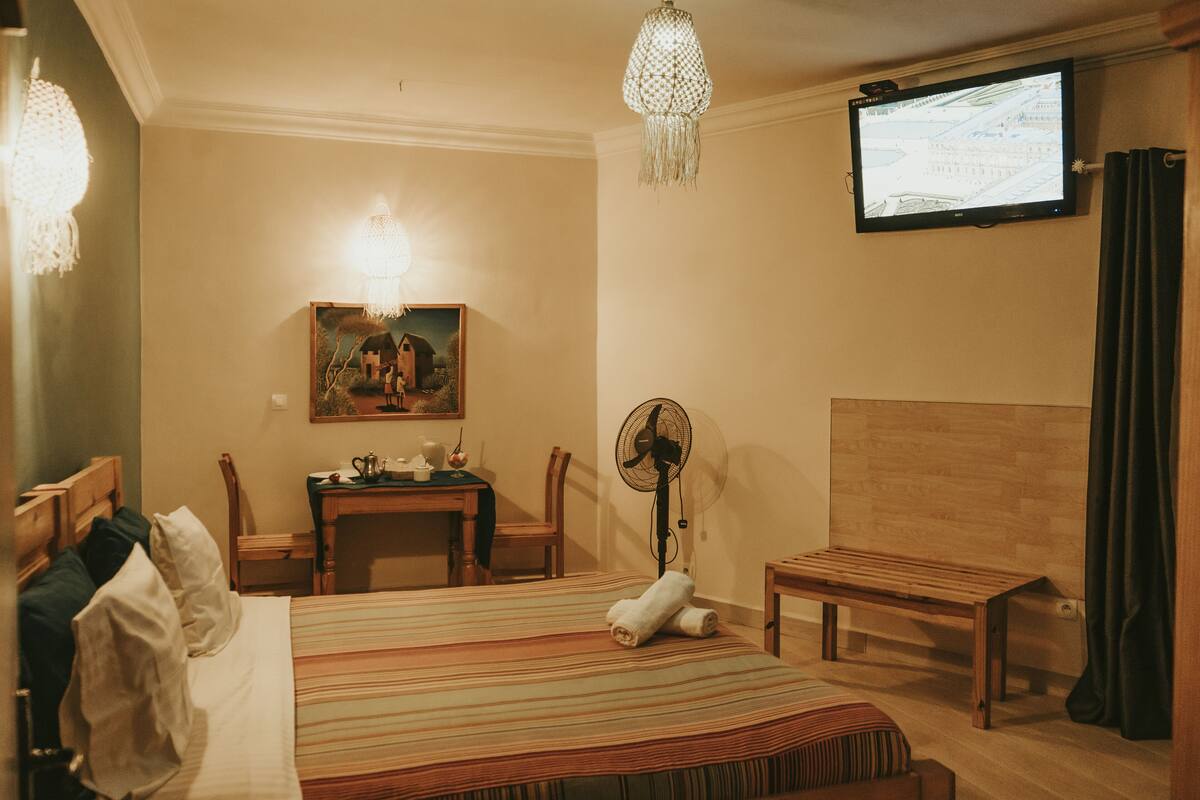 A hotel room with a bed, a desk, a chair, a fan, a television, and a painting on the wall.