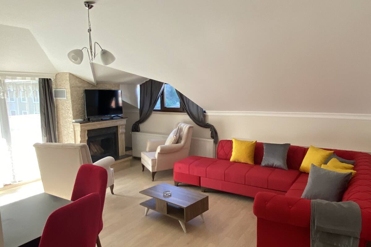 Amplio salón con un sofá rojo, un sillón blanco, una butaca roja, una mesa de centro y un televisor de pantalla plana montado en la pared.
