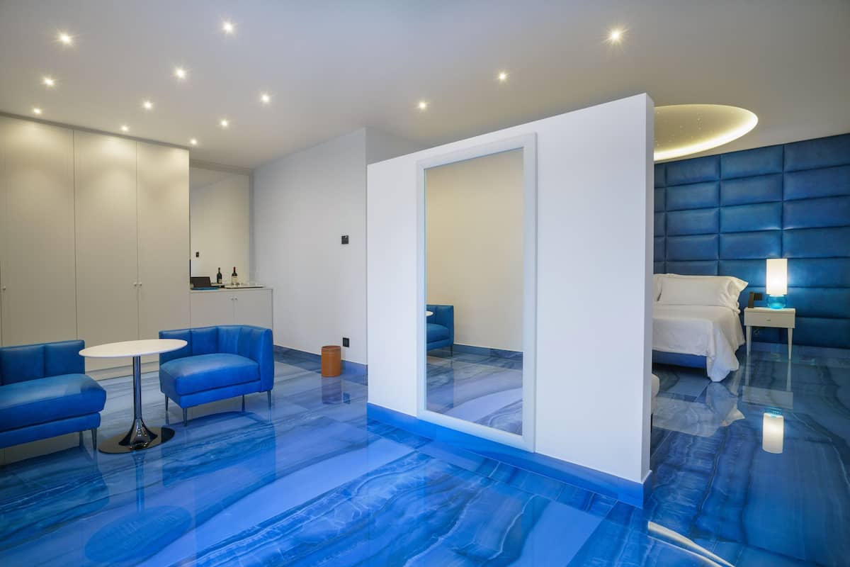 Una habitación de hotel moderna con piso de mármol azul, una cama blanca, un sofá azul y una mesita pequeña.
