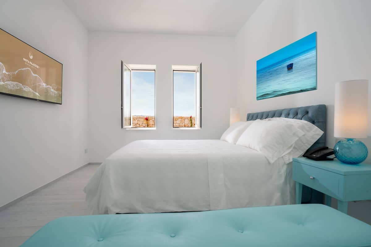 Un dormitorio con una cama, una mesita de noche, una lámpara y dos ventanas.