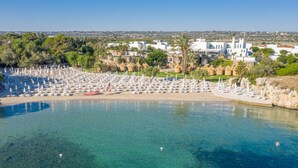 A coastal resort with clear blue waters, numerous beach umbrellas, and a backdrop of white buildings and greenery.