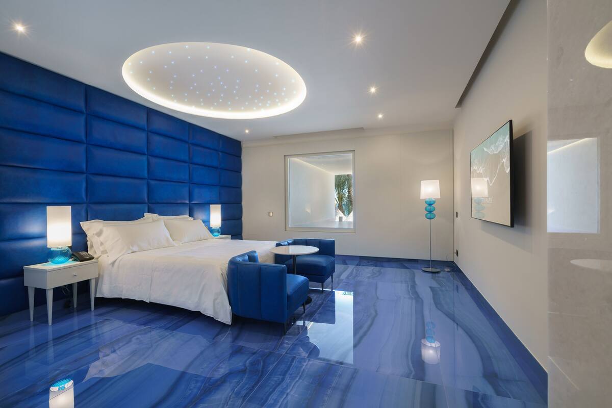 Un dormitorio moderno con una pared azul de acento, una cama grande, una mesita de noche con lámpara, una pequeña mesa redonda y un espejo.