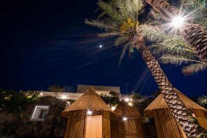 A night scene with a palm tree, thatched-roof structures, and a building in the background.