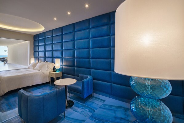 A modern hotel room with a blue padded headboard, a white bed, a blue sofa, a round table, and a unique blue glass lamp. - Grotta Palazzese Beach Hotel (Polignano a Mare)