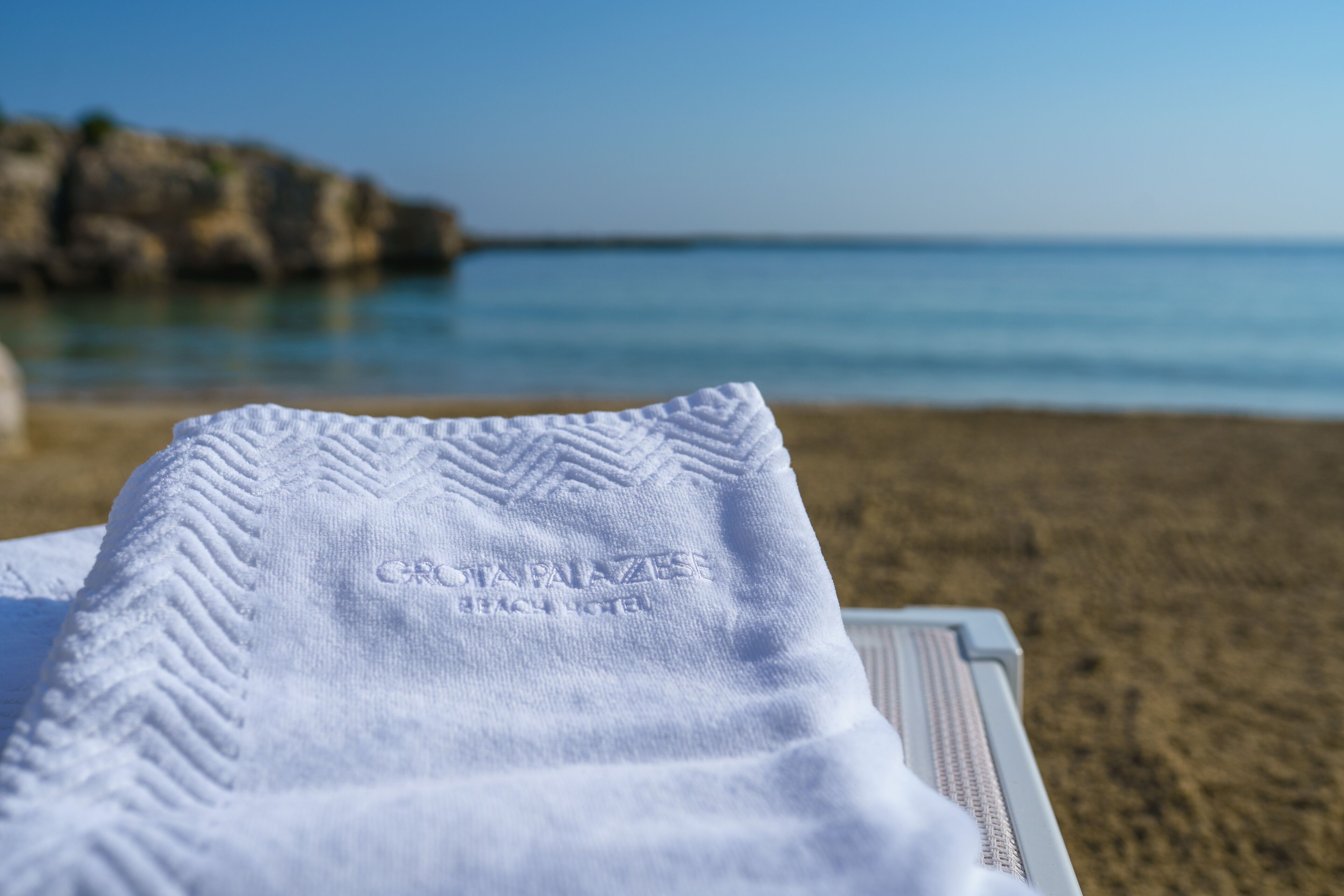 Una toalla blanca con el texto grabado 'GRIAZZA BEACH HOTEL', sobre una mesa con vista a una playa y un litoral rocoso.
