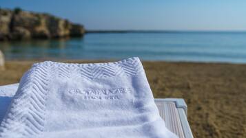 Una toalla blanca con el texto grabado 'GRIAZZA BEACH HOTEL', sobre una mesa con vista a una playa y un litoral rocoso.