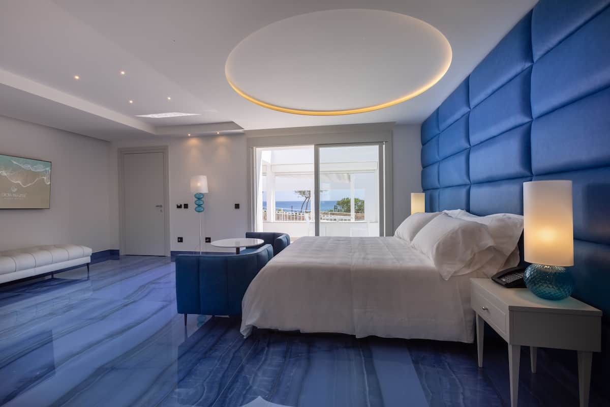 Una habitación de hotel moderna con una cama grande, un escritorio con lámpara, una silla azul y vista al mar a través de una puerta corrediza.