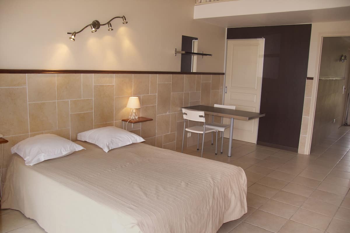 A hotel room with a bed, a desk, a chair, a lamp, and a door leading to another room.