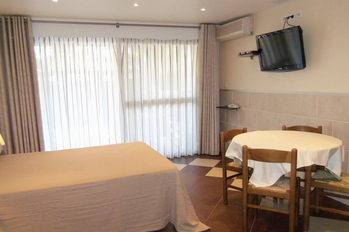 A hotel room with a bed, a table with chairs, a TV mounted on the wall, and a window with curtains.
