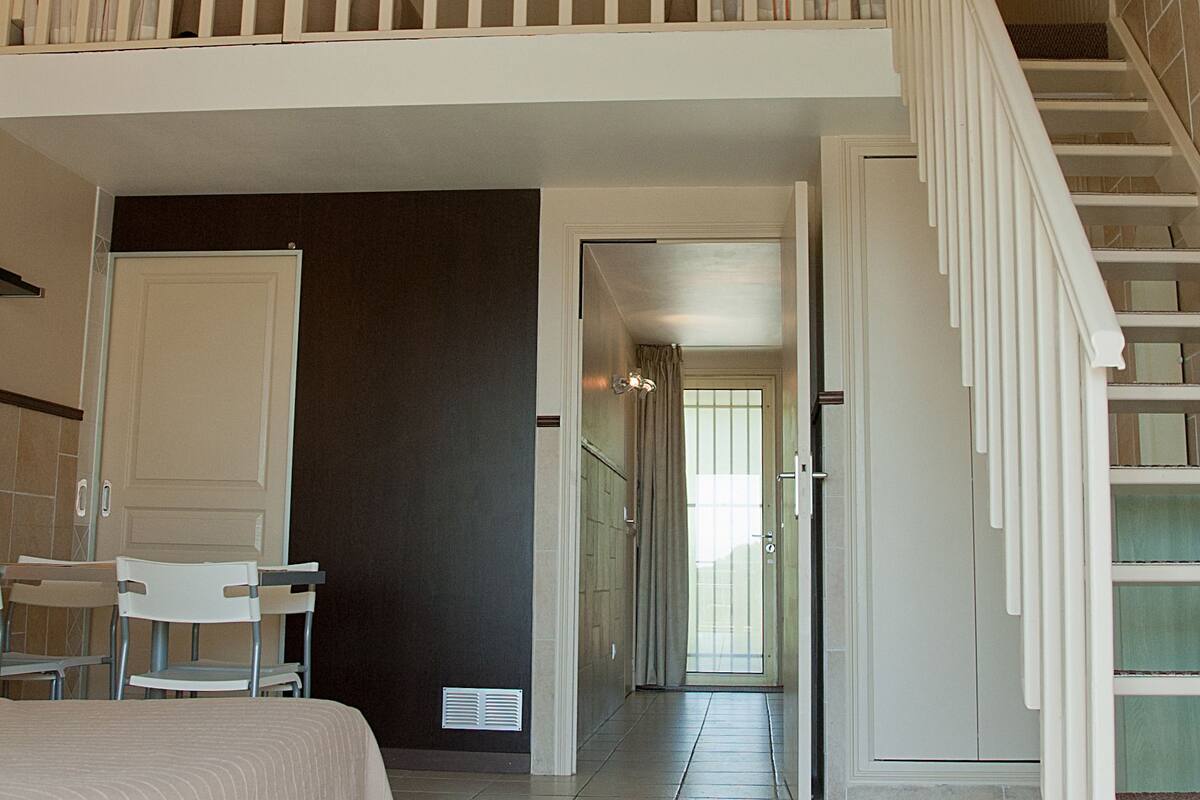 A spacious room with a high ceiling, a staircase, and a view into another room.