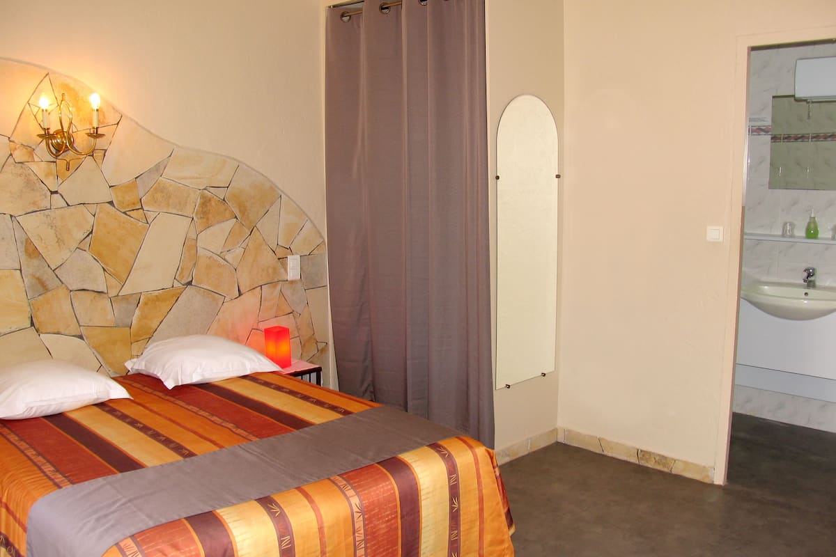 A bedroom with a stone headboard, a bed with striped bedding, a nightstand with a lamp, and a bathroom with a sink and mirror.