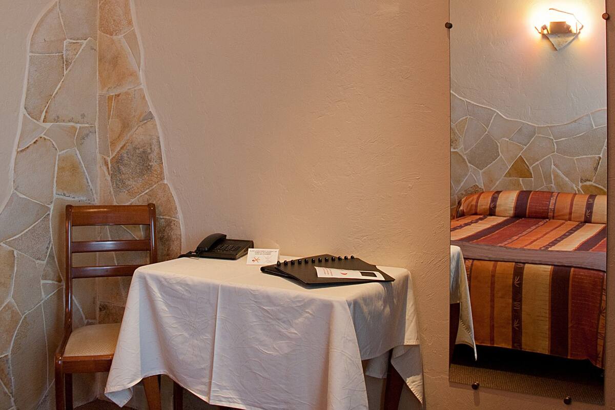 A hotel room with a stone wall, a flat-screen TV mounted above a table with a white tablecloth, a wooden chair, and a bed with a striped blanket.