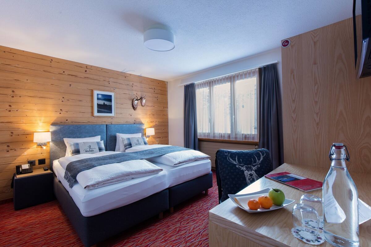 Ein Hotelzimmer mit Holzwand, einem Bett mit Kopfteil, einem Nachttisch, einem Schreibtisch mit Obstteller, einer Flasche und einem Stuhl.