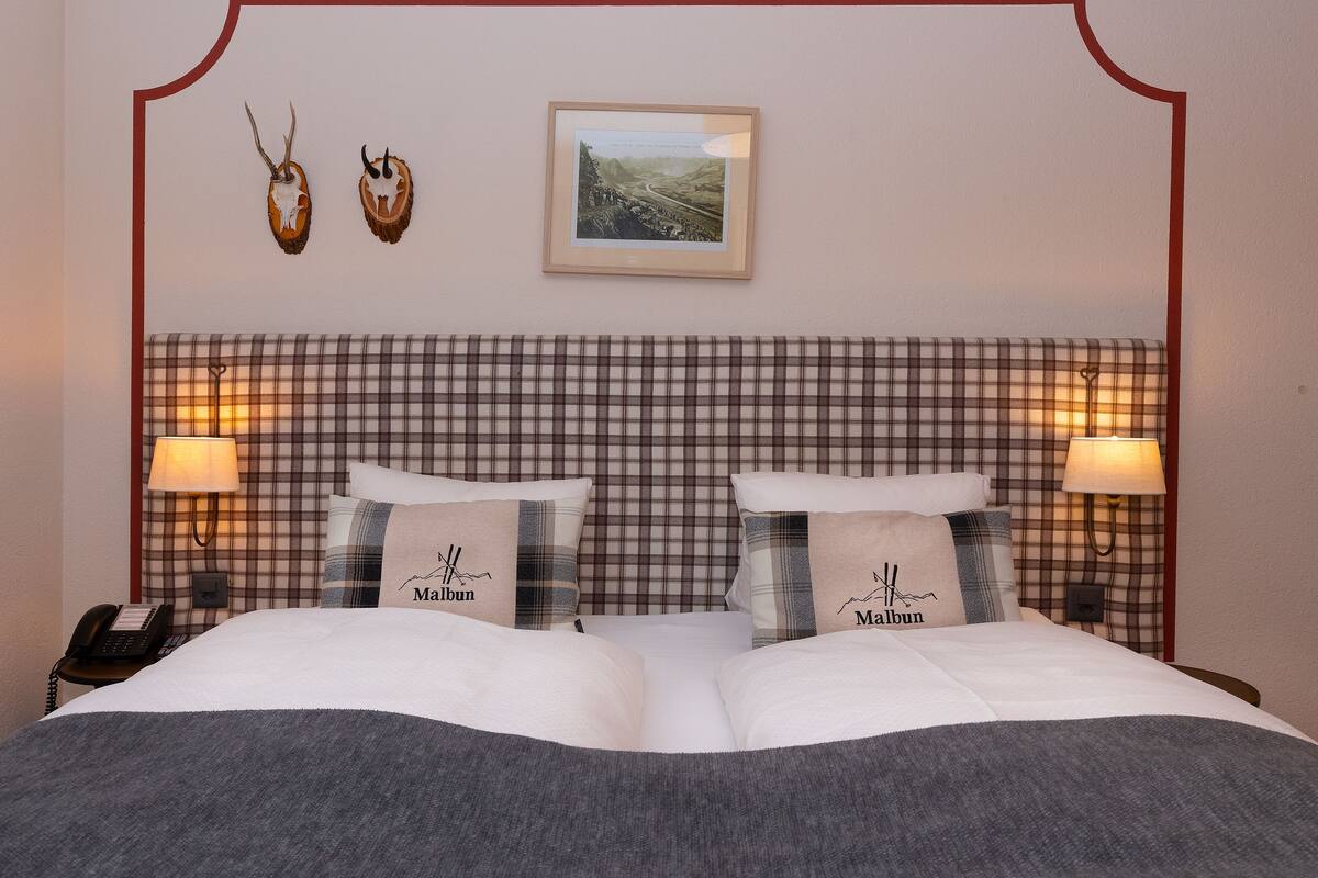 Ein Hotelzimmer mit einem Karo-Kopfstütze, zwei Nachttischlampen, einem gerahmten Bild und zwei Kissen mit dem Schriftzug 'Malbun'.