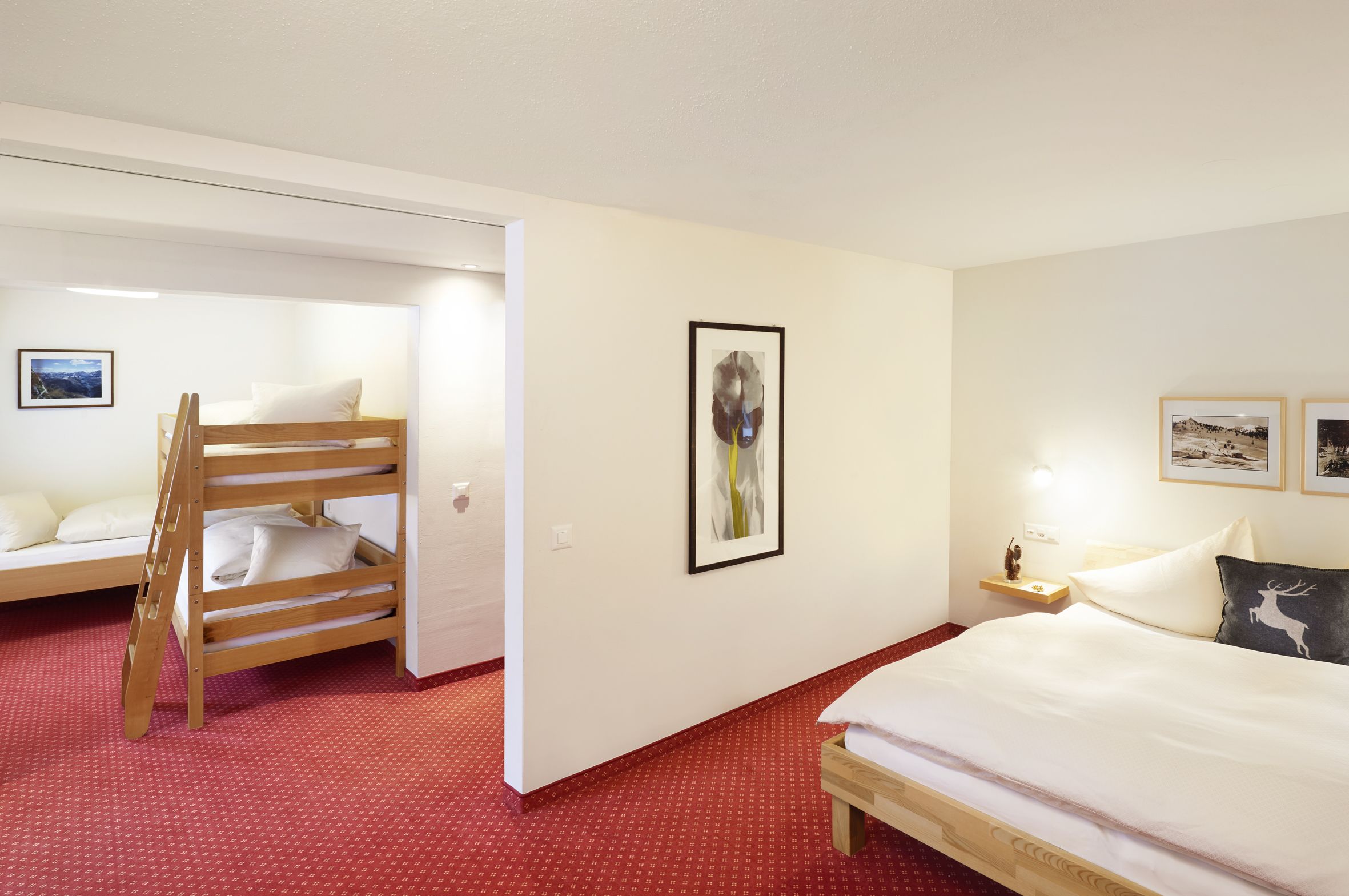 A hotel room with a bunk bed, a single bed, a small bedside table with a lamp, and a framed picture on the wall.