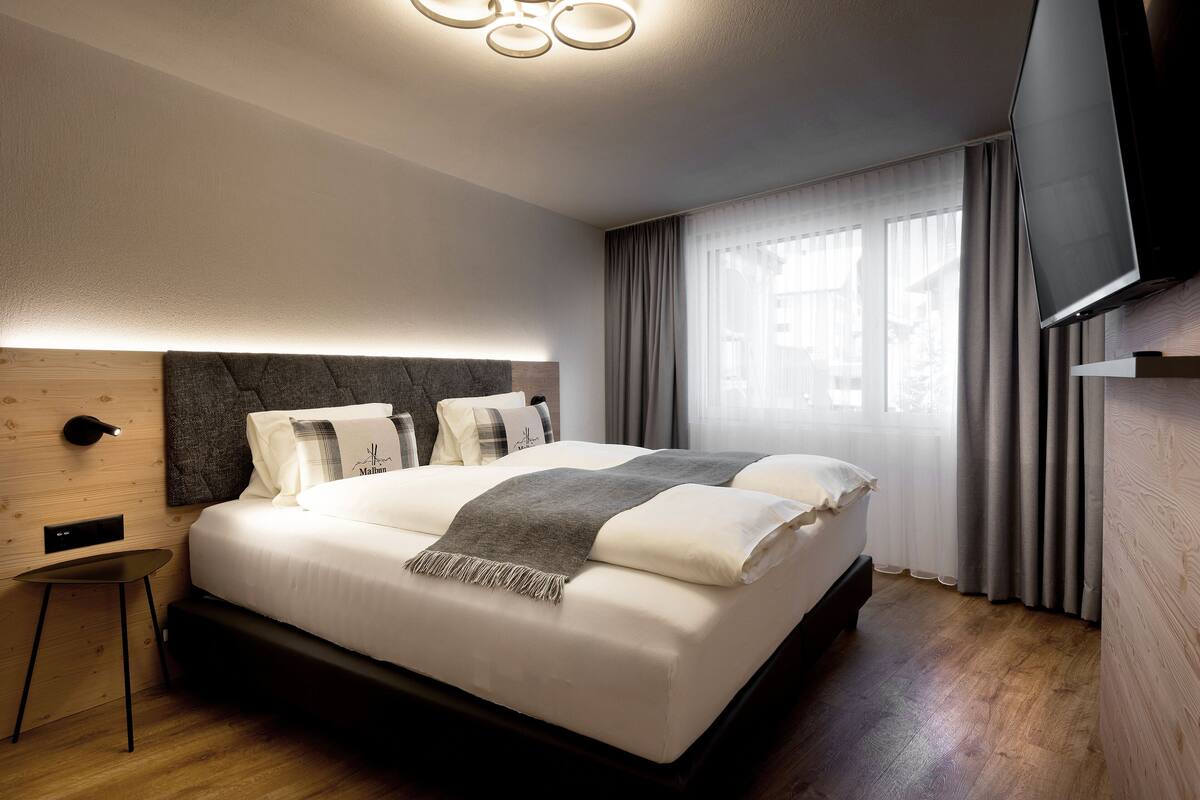 Ein modernes Hotelzimmer mit einem großen Bett, einem kleinen Nachttisch, einem Flachbildfernseher und einem großen Fenster mit Vorhängen.