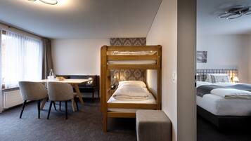 Una habitación de hotel con una cama litera, un escritorio con sillas, una mesita pequeña y un banco.