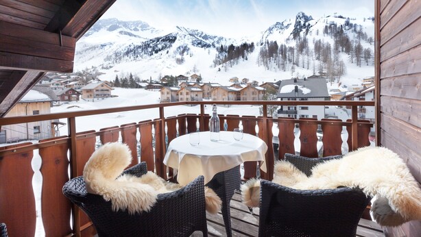 Ein Holzbalkon mit Korbstühlen und einem Tisch, mit Blick auf ein verschneites Bergdorf.