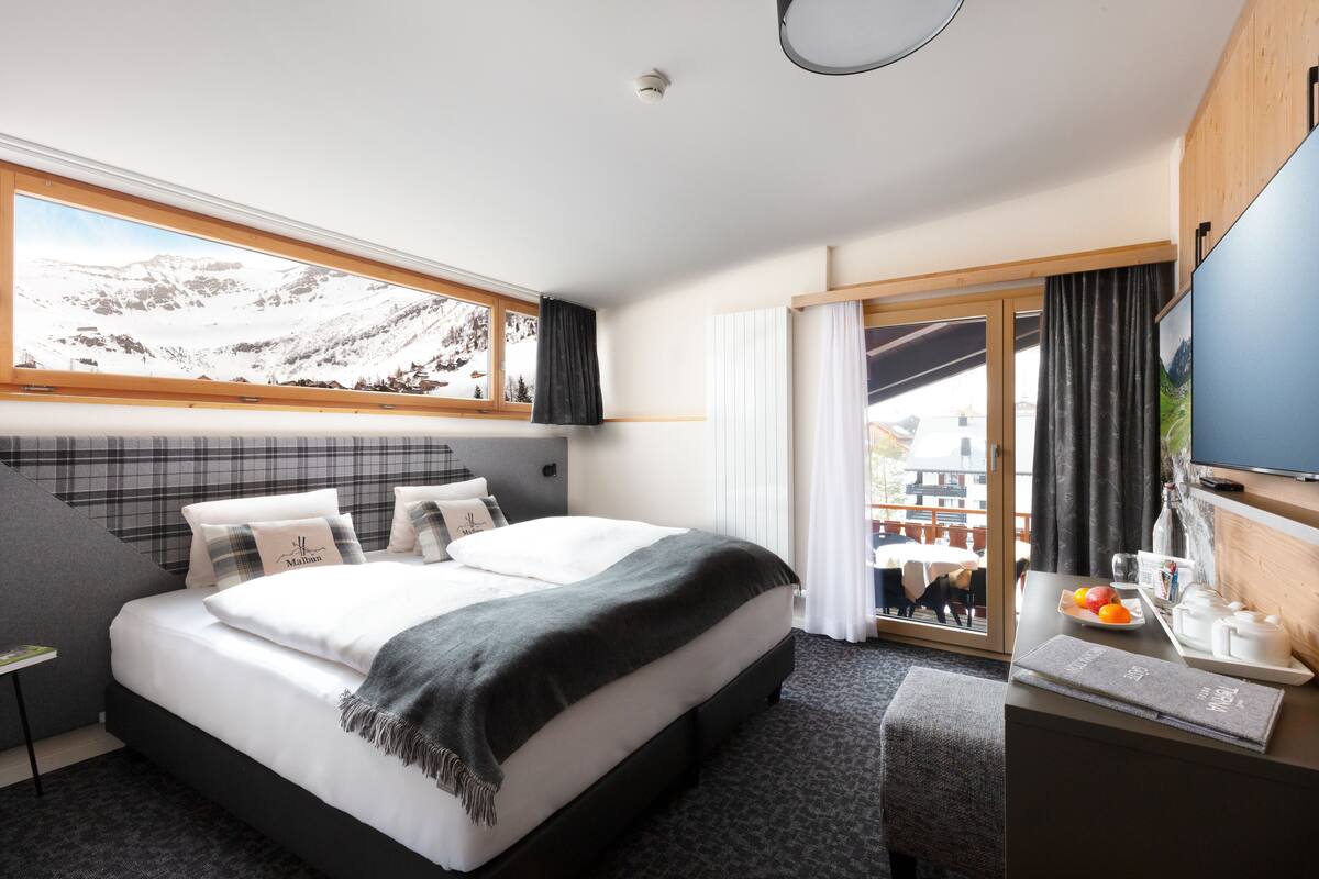 Ein modernes Hotelzimmer mit einem großen Bett, einem Schreibtisch mit einem Laptop, einem Fernseher und Blick auf Berge durch das Fenster.