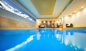 Ein Swimmingpool mit einer Bergmalerei an der Wand, ein Empfangsbereich mit einer Couch und einem Holzkabinent, und eine Glastür, die zu einem Raum führt.