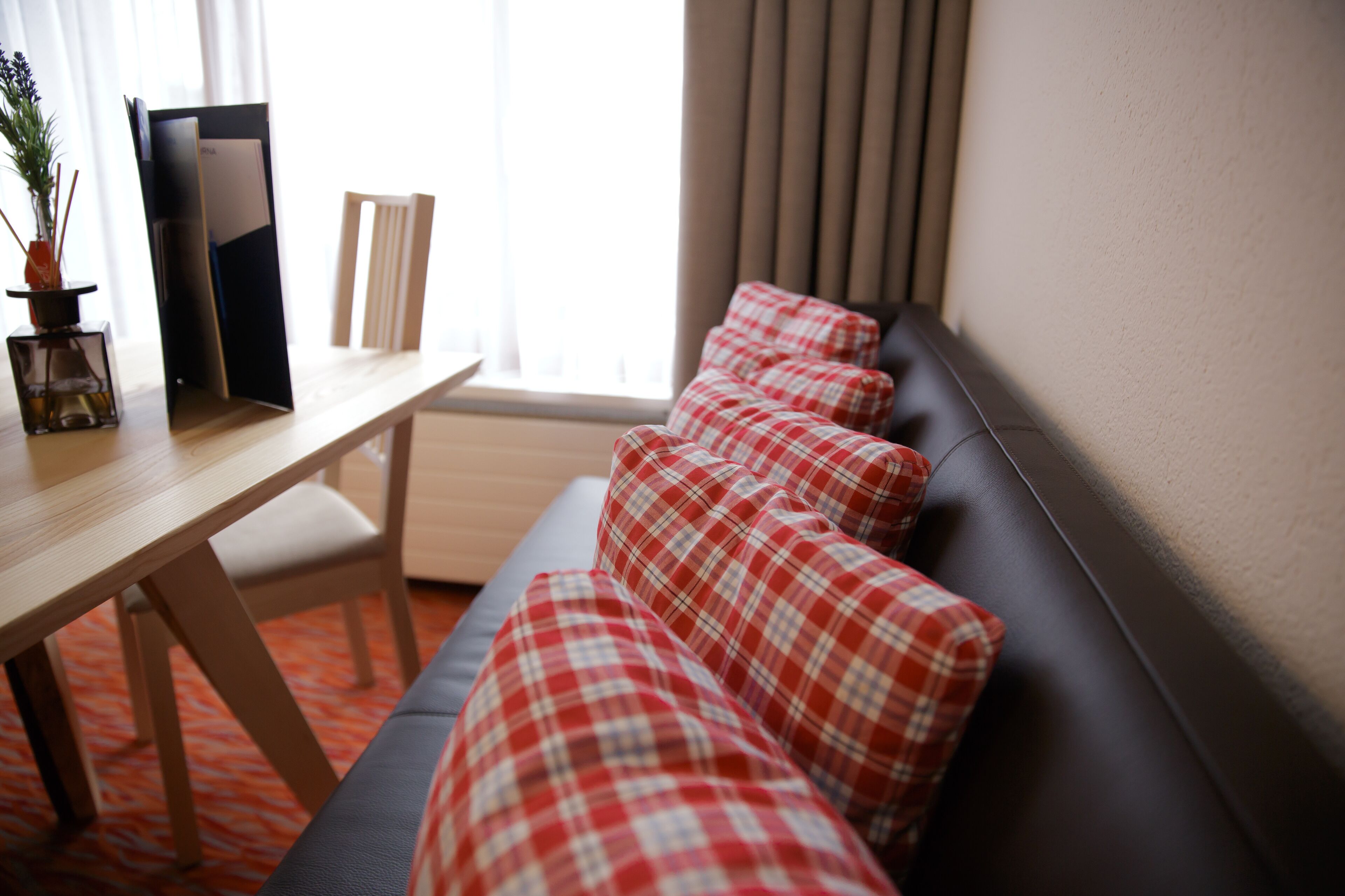 Una habitación con una mesa de madera, un sofá negro con cojines a cuadros rojos y blancos, y una ventana con persianas verticales.